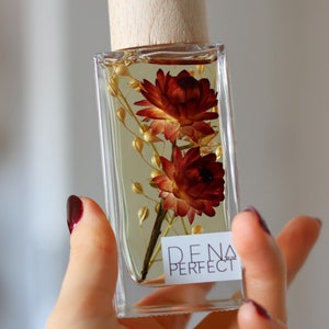 Può includere: Una bottiglia di vetro trasparente con un tappo di legno e fiori secchi all'interno. La bottiglia è etichettata "DENA PERFECT".