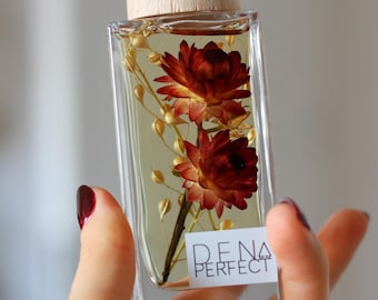 Puede incluir: Una botella de vidrio transparente con tapa de madera y flores secas en su interior. La botella está etiquetada con "DENA PERFECT".