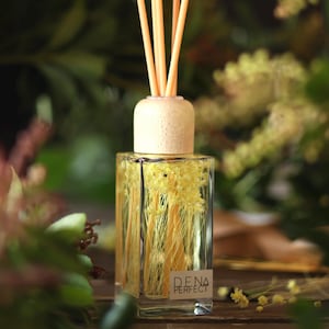Può includere: Una bottiglia di diffusore in vetro trasparente con un tappo in legno e canne naturali. La bottiglia è riempita con un liquido giallo chiaro e fiori secchi. La bottiglia è etichettata "DENA PERFECT".