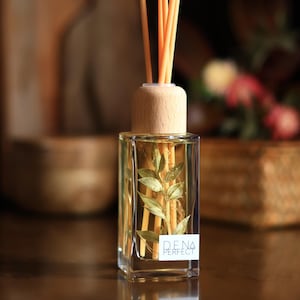 Könnte beinhalten: Ein klarer Glasdiffusor mit einem Holzdeckel und orangefarbenen Stäbchen. Die Flasche enthält eine gelbe Flüssigkeit und dekorative Blätter. Auf dem Etikett steht "DENA PERFECT". Der Hintergrund ist verschwommen, mit einer Holzschale und Blumen.