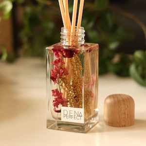 Könnte beinhalten: Eine klare, rechteckige Diffusorflasche mit getrockneten Blumen und Stäbchen. Die Flasche hat einen Holzdeckel daneben. Auf dem Etikett der Flasche steht "D.E.N.A. PERFECT". Der Hintergrund ist mit grünem Laub verschwommen.