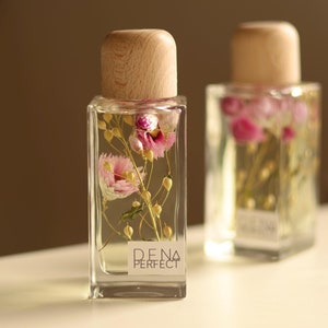 Può includere: Due bottiglie di vetro trasparente con tappi di legno riempite di fiori secchi e un liquido trasparente. La bottiglia a sinistra ha un'etichetta con la scritta "D.E.N.A PERFECT".