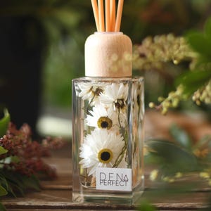 May include: A clear glass diffuser bottle with a wooden lid and reeds. The bottle contains white flowers and a label that reads "DENA PERFECT".