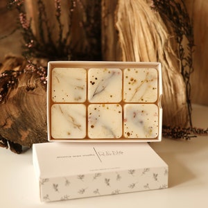 Puede incluir: Una caja blanca con seis pequeñas pastillas de cera fundente cuadradas en su interior. Las pastillas de cera son blancas con un patrón de mármol y motas doradas. La caja está etiquetada con "aroma wax melts" y "Peter & Lily".