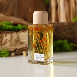 May include: A clear glass bottle with a wooden lid filled with a yellow liquid and dried flowers. The bottle is labelled "DENA PERFECT".