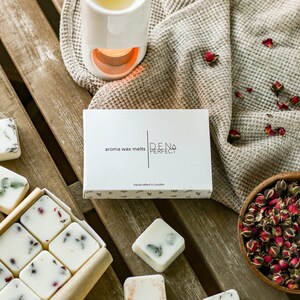 Puede incluir: Una caja de fundidos de cera blanca con el texto "aroma wax melts DENA PERFECT handcrafted in London" en la parte delantera. La caja est&aacute; sobre una superficie de madera con un calentador de cera de cer&aacute;mica blanca y un cuenco de p&eacute;talos de rosa secos.