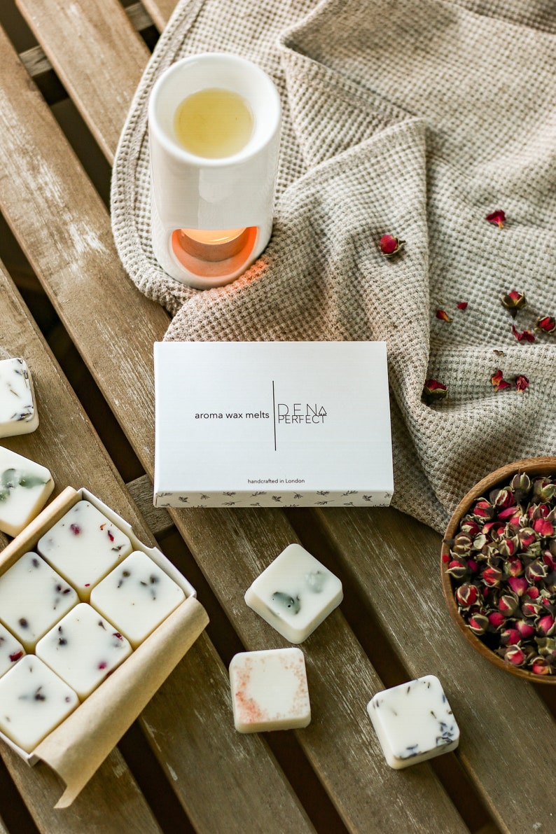Puede incluir: Una caja de fundidos de cera blanca con el texto "aroma wax melts DENA PERFECT handcrafted in London" en la parte delantera. La caja est&aacute; sobre una superficie de madera con un calentador de cera de cer&aacute;mica blanca y un cuenco de p&eacute;talos de rosa secos.