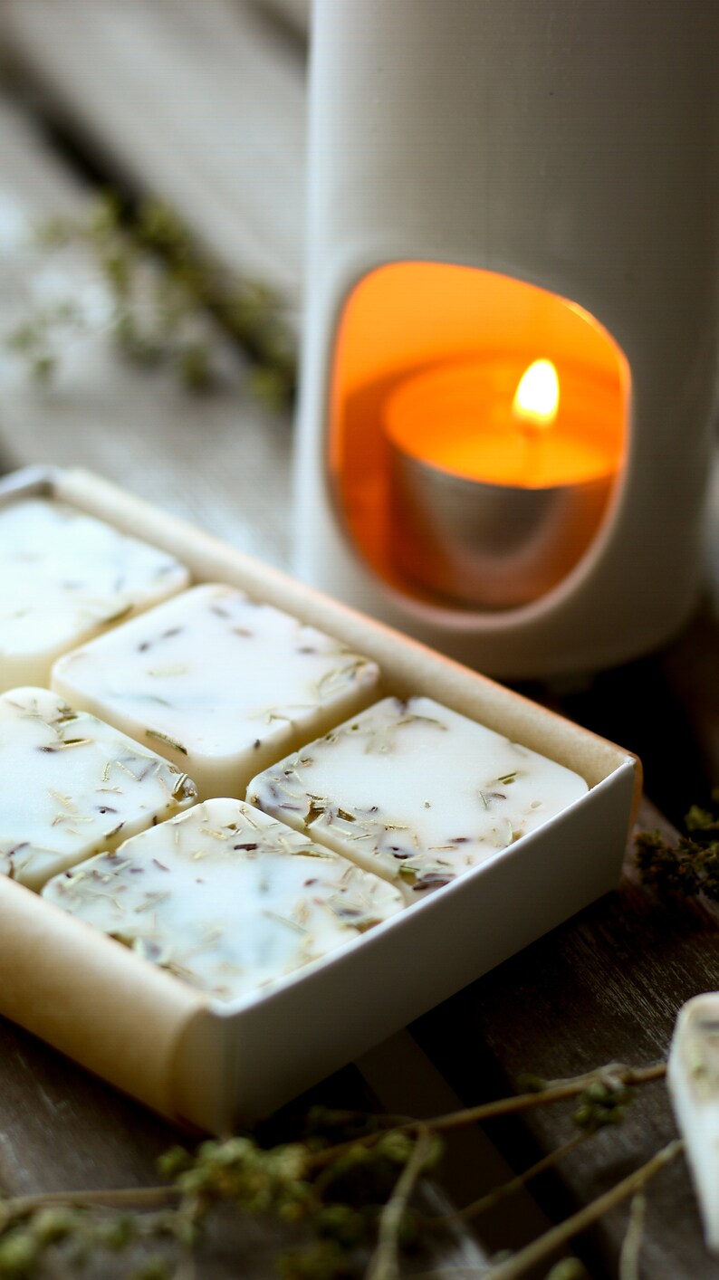 Puede incluir: Una caja blanca llena de seis cubos de cera derretida, cada uno con hierbas y flores secas. La caja est&aacute; sobre una superficie de madera junto a un calentador de velas de cer&aacute;mica blanca con una vela de t&eacute; encendida dentro.