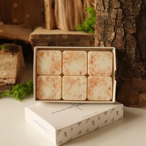 May include: A white box with a floral pattern containing six square wax melts. The wax melts are a light beige colour with a glittery finish. The box says "aroma wax melts" and "handcrafted in London".