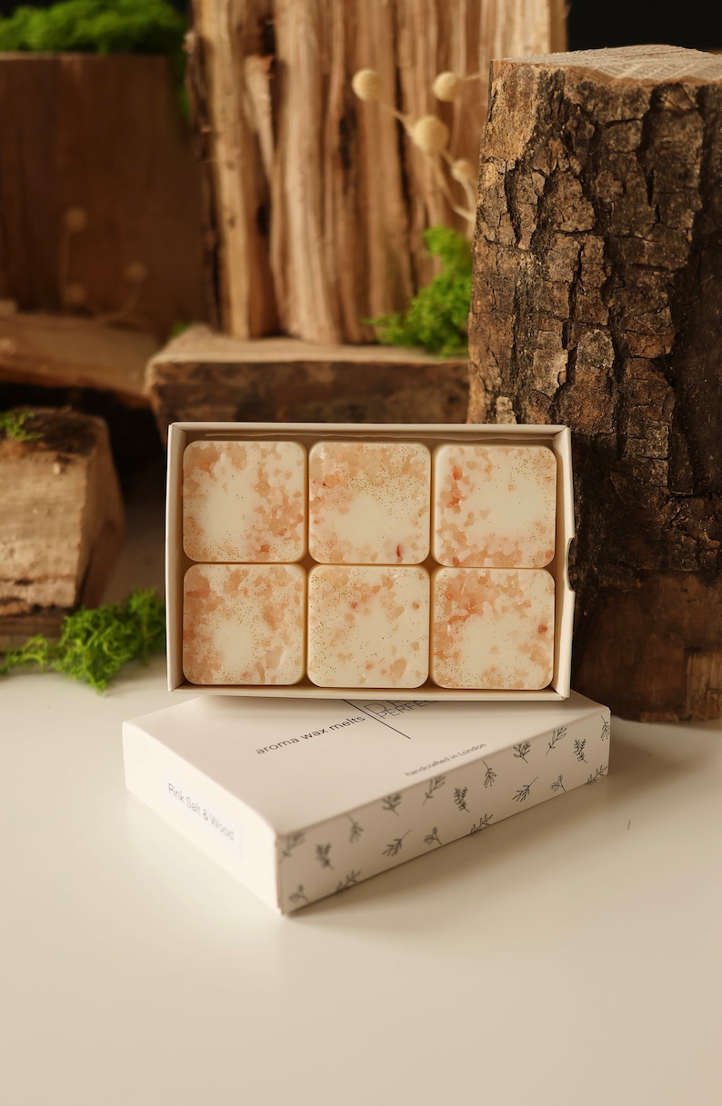 K&ouml;nnte beinhalten: Eine wei&szlig;e Schachtel mit Blumenmuster, die sechs quadratische Wachs-Schmelzen enth&auml;lt. Die Wachs-Schmelzen sind hellbeige mit einem glitzernden Finish. Die Schachtel tr&auml;gt die Aufschrift "aroma wax melts" und "handcrafted in London".