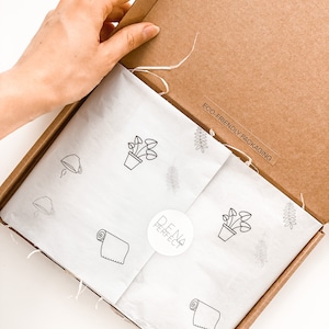 Puede incluir: Caja de cart&oacute;n abierta con papel de seda blanco en el interior. El papel de seda tiene un patr&oacute;n repetido de una planta en maceta, una toalla enrollada y una hoja. La caja tiene una pegatina que dice "EMBALAJE ECOL&Oacute;GICO" y una pegatina con el texto "DENA PERFECT".