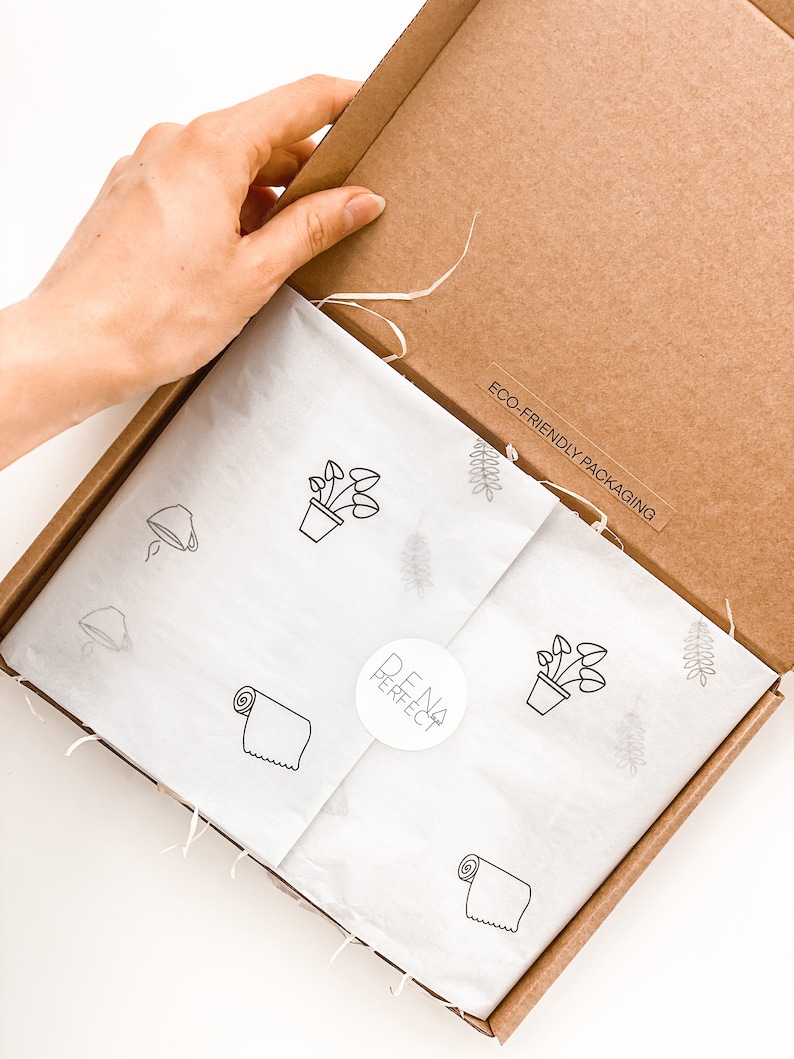 Puede incluir: Caja de cart&oacute;n abierta con papel de seda blanco en el interior. El papel de seda tiene un patr&oacute;n repetido de una planta en maceta, una toalla enrollada y una hoja. La caja tiene una pegatina que dice "EMBALAJE ECOL&Oacute;GICO" y una pegatina con el texto "DENA PERFECT".