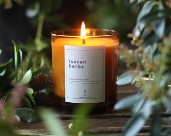 May include: A brown glass candle with a white label that reads "tuscan herbs handcrafted soy candle". The candle is lit and the flame is visible.