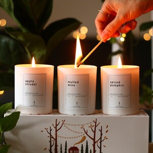 Puede incluir: Tres velas blancas con etiquetas que dicen "apple strudel", "vino caliente" y "calabaza especiada". Las velas est&aacute;n dispuestas en una caja blanca con una escena de bosque y el texto "Colecci&oacute;n de velas de oto&ntilde;o de Den Perfect Hecho a mano en Londres".