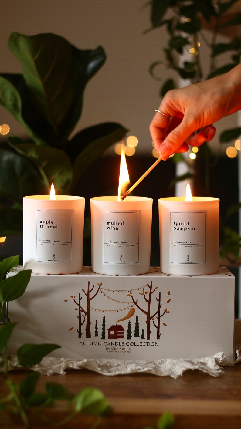 Puede incluir: Tres velas blancas con etiquetas que dicen "apple strudel", "vino caliente" y "calabaza especiada". Las velas est&aacute;n dispuestas en una caja blanca con una escena de bosque y el texto "Colecci&oacute;n de velas de oto&ntilde;o de Den Perfect Hecho a mano en Londres".