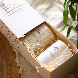 Puede incluir: Una caja de regalo blanca con un dise&ntilde;o de oto&ntilde;o en la tapa. La caja est&aacute; abierta y contiene dos velas blancas con etiquetas que dicen "Spiced Pumpkin" y "Mulled Wine". Las velas est&aacute;n anidadas en papel triturado.