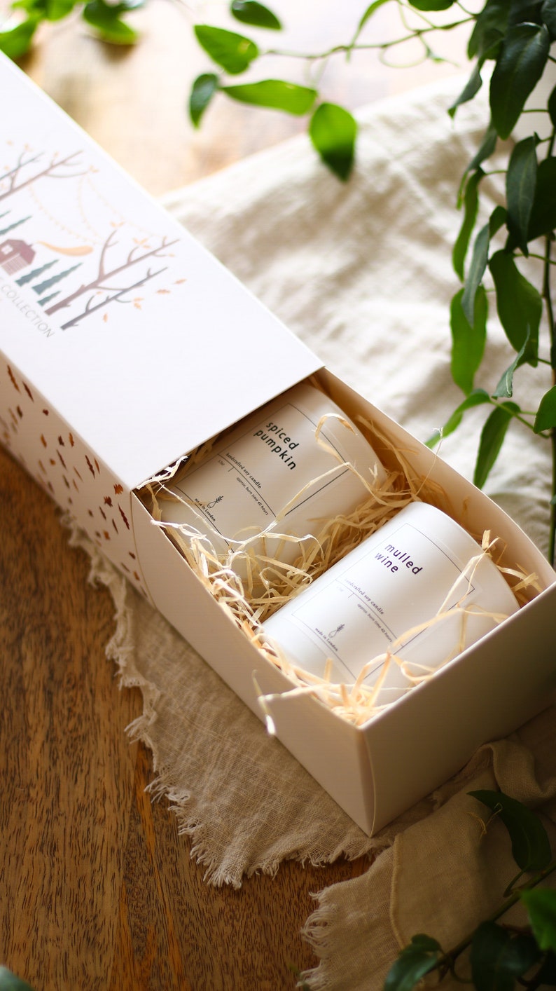 Puede incluir: Una caja de regalo blanca con un dise&ntilde;o de oto&ntilde;o en la tapa. La caja est&aacute; abierta y contiene dos velas blancas con etiquetas que dicen "Spiced Pumpkin" y "Mulled Wine". Las velas est&aacute;n anidadas en papel triturado.