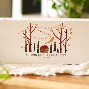 Puede incluir: Una caja blanca con una ilustraci&oacute;n marr&oacute;n y naranja de una caba&ntilde;a en un bosque con &aacute;rboles y luces de cadena. El texto en la caja dice "Autumn Candle Collection by Den Perfect Handmade in London."