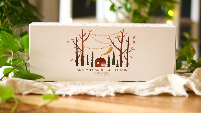 Puede incluir: Una caja blanca con una ilustraci&oacute;n marr&oacute;n y naranja de una caba&ntilde;a en un bosque con &aacute;rboles y luces de cadena. El texto en la caja dice "Autumn Candle Collection by Den Perfect Handmade in London."