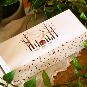 Puede incluir: Una caja blanca con una ilustraci&oacute;n marr&oacute;n y dorada de una caba&ntilde;a en el bosque con una cadena de luces. El texto en la caja dice "Autumn Candle Collection by Den Perfect Handmade in London". La caja est&aacute; rodeada de hojas verdes.