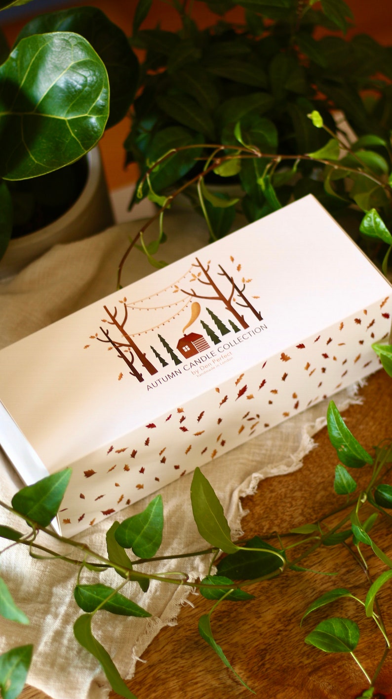 Puede incluir: Una caja blanca con una ilustraci&oacute;n marr&oacute;n y dorada de una caba&ntilde;a en el bosque con una cadena de luces. El texto en la caja dice "Autumn Candle Collection by Den Perfect Handmade in London". La caja est&aacute; rodeada de hojas verdes.