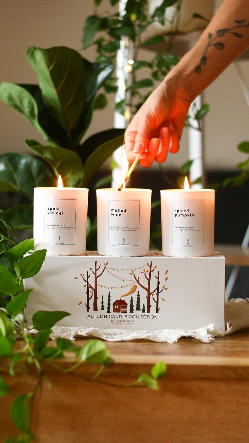 Puede incluir: Tres velas blancas con etiquetas que dicen "apple strudel", "vino caliente" y "calabaza especiada". Las velas est&aacute;n dispuestas en una caja blanca con una escena de bosque y el texto "Colecci&oacute;n de velas de oto&ntilde;o de Den Perfect Hecho a mano en Londres".