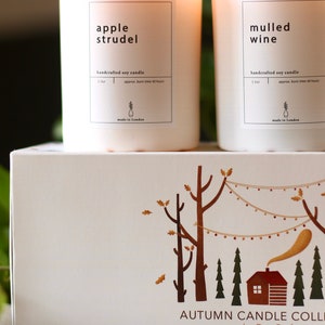 Puede incluir: Dos velas de soja blancas con etiquetas que dicen "apple strudel" y "vino caliente". Las velas son artesanales y tienen un tiempo de combusti&oacute;n aproximado de 40 horas. Las velas est&aacute;n sobre una caja blanca con una ilustraci&oacute;n marr&oacute;n de una caba&ntilde;a en el bosque. La caja dice "Colecci&oacute;n de Velas de Oto&ntilde;o por Den Perfect, hecha a mano en Londres."