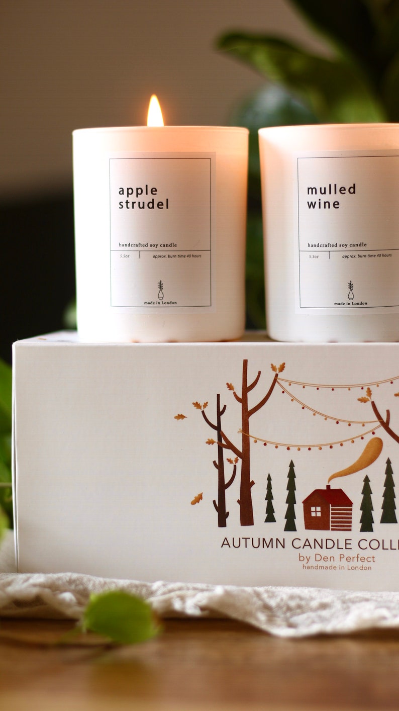 Puede incluir: Dos velas de soja blancas con etiquetas que dicen "apple strudel" y "vino caliente". Las velas son artesanales y tienen un tiempo de combusti&oacute;n aproximado de 40 horas. Las velas est&aacute;n sobre una caja blanca con una ilustraci&oacute;n marr&oacute;n de una caba&ntilde;a en el bosque. La caja dice "Colecci&oacute;n de Velas de Oto&ntilde;o por Den Perfect, hecha a mano en Londres."