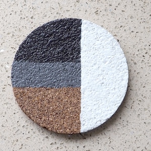 Puede incluir: Un posavasos redondo de corcho con un diseño geométrico. El posavasos está pintado en cuatro secciones: negro, gris, marrón y blanco.