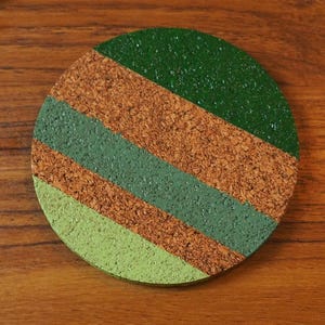 May include: Circular cork coaster featuring diagonal stripes in green and brown. The coaster is made from natural cork and has a painted design. A decorative and practical item for home use, measuring approximately 10 cm in diameter.