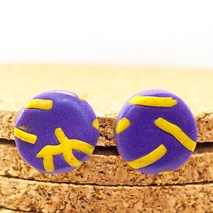 May include: A pair of purple and yellow stud earrings with a geometric pattern. The earrings are round and have a shiny finish.