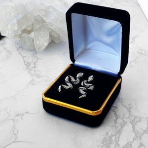 May include: A pair of silver snake earrings in a black velvet jewelry box with a gold trim.