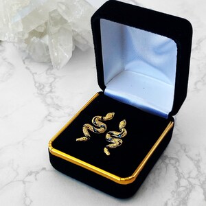 May include: A pair of gold snake earrings with blue accents, displayed in a black velvet jewelry box with a gold trim.