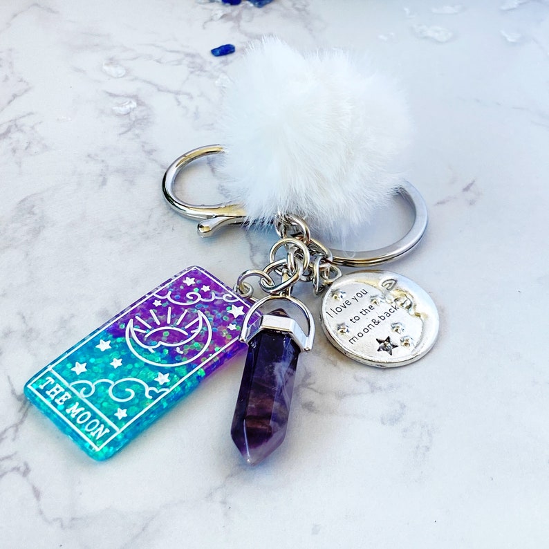 Peut inclure: Un porte-cl&eacute;s argent&eacute; avec un charme en cristal violet, un pompon blanc et un charme argent&eacute; avec le texte "Je t'aime jusqu'&agrave; la lune et retour". Le porte-cl&eacute;s comprend &eacute;galement une carte bleue et violette avec des paillettes et le texte "La Lune".