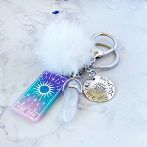 Peut inclure: Un porte-cl&eacute;s argent&eacute; avec un charme pompon blanc, un charme en cristal transparent et un charme argent&eacute; qui dit "You are my sunshine". Le porte-cl&eacute;s a &eacute;galement une carte avec un soleil et les mots "The Sun" dessus.