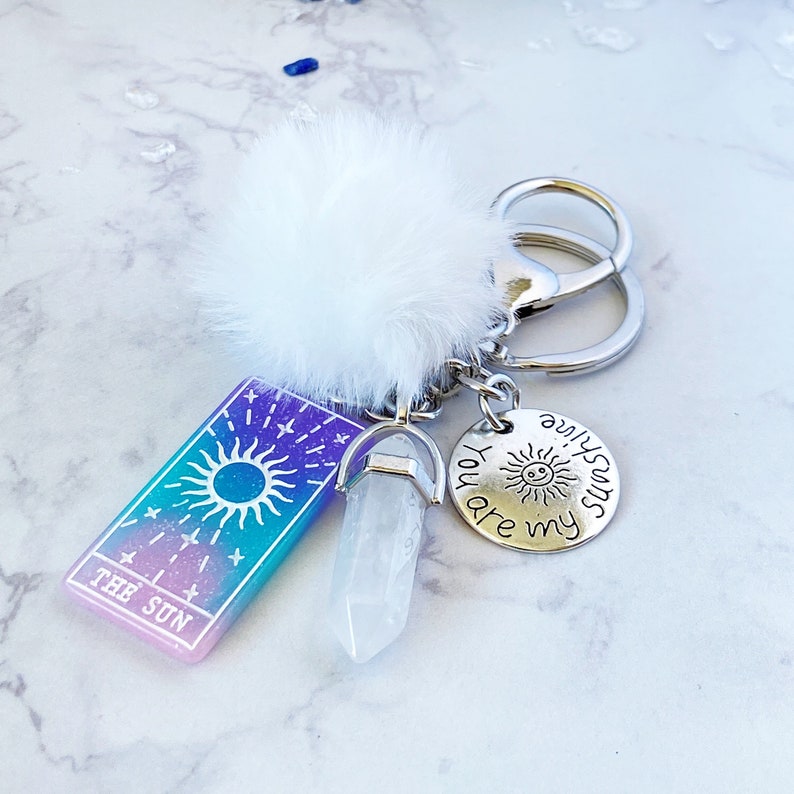 Peut inclure: Un porte-cl&eacute;s argent&eacute; avec un charme pompon blanc, un charme en cristal transparent et un charme argent&eacute; qui dit "You are my sunshine". Le porte-cl&eacute;s a &eacute;galement une carte avec un soleil et les mots "The Sun" dessus.