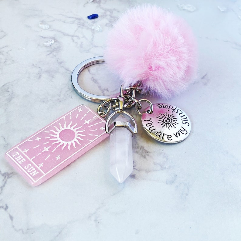 Peut inclure: Porte-cl&eacute;s rose et argent avec un pompon rose, un charme en cristal transparent et un charme argent&eacute; qui dit "You are my sunshine". Le charme rose pr&eacute;sente un soleil et des &eacute;toiles et le texte "The Sun".