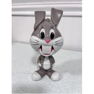 May include: A gray and white plastic rabbit figurine featuring large ears, black oval eyes, a small pink nose, and prominent front teeth. The character has a rounded head and body with white accents on the face, chest, and feet, standing upright.