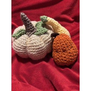 May include: A crocheted off-white pumpkin with a dark grey stem and green leaves, accompanied by a crocheted orange pear with a brown stem and a light yellow stem-like extension, all crafted with visible yarn texture and intricate stitching.
