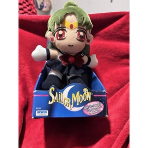 May include: A Sailor Moon plush toy with green hair, red eyes, and a yellow tiara with a red gem. The character wears a dark blue sailor-style uniform with a red bow. It is packaged in a blue box labelled Sailor Moon, Soft 'n Cuddly, and Irwin.