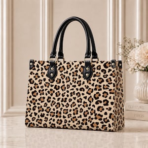 May include: A structured rectangular handbag featuring a tan and black leopard print pattern. The bag includes two black top handles attached with gold-toned metal hardware and rivets. The design has a flat base and vertical side panels.
