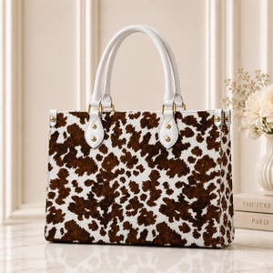 Puede incluir: Un bolso estructurado con un patrón de estampado de vaca en marrón y blanco. El bolso tiene dos asas superiores blancas sujetas con herrajes dorados, incluyendo tachuelas decorativas en la base de cada asa y en los paneles laterales del bolso.