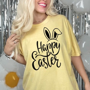 Könnte beinhalten: Ein gelbes T-Shirt mit einer schwarzen Grafik, auf der "Happy Easter" mit Hasenohren über dem "H" steht.