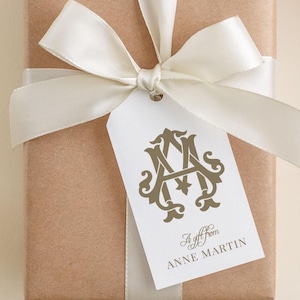 May include: A rectangular gift wrapped in brown paper, tied with a white satin ribbon and bow. A white tag with gold lettering and a monogram is attached to the bow. The tag reads "A gift from Anne Martin."