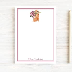 May include: A white notepad with a pink border and a watercolor illustration of a golden retriever holding flowers. The notepad has the name "Claire Anderson" printed at the bottom. A pencil rests on the right side.