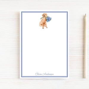 May include: A white notepad with a blue border and a watercolor illustration of a golden retriever holding blue flowers. A pencil rests to the right. The name "Claire Anderson" is printed at the bottom.