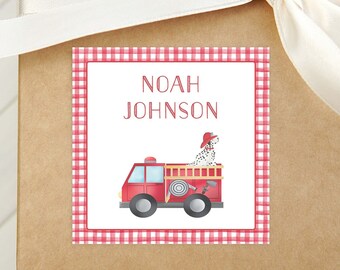Op de afbeelding: Een vierkant cadeaulabel met een rode ruitjesrand en de naam "NOAH JOHNSON" in rode letters. Daaronder een rode brandweerwagen met een Dalmatische hond met een brandweerhoed. Het label zit op een bruine geschenkdoos met een wit lint.