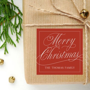 May include: A wrapped gift with a red square label that reads "Merry Christmas" in elegant white script, with "The Thomas Family" below. The package is tied with twine and adorned with a small gold bell. Green sprigs and additional gold bells are visible.