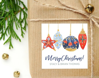 Può includere: Un regalo confezionato con un'etichetta quadrata con decorazioni natalizie ad acquerello. L'etichetta recita "Merry Christmas!" con un nome. Il regalo è legato con spago e decorato con una campana dorata e rametti di sempreverdi.