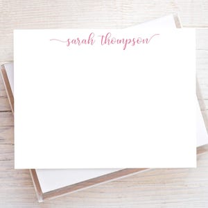 May include: White stationery with the name "sarah thompson" in a cursive, pink font. The stationery is stacked in a clear box, resting on a light wood surface. The design is simple and elegant.